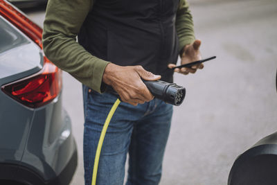 Midsection of mature man holding smart phone and electric charger plug at station