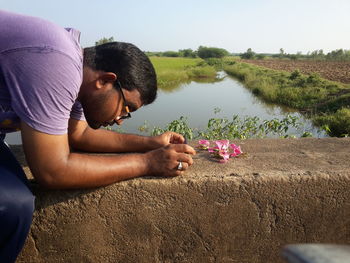 Man photographing flower while sitting on retaining wall against sky