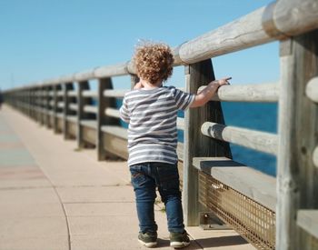 Rear view of boy walking on footbridge over sea against sky