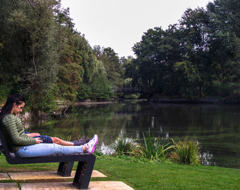 Side view of woman sitting by lake against trees