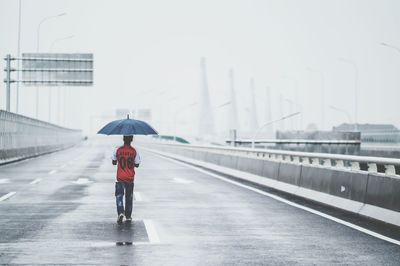 Rear view of man carrying umbrella while walking on road during rainy season