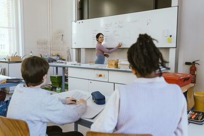 Smiling female teacher writing on whiteboard while teaching male students sitting in classroom