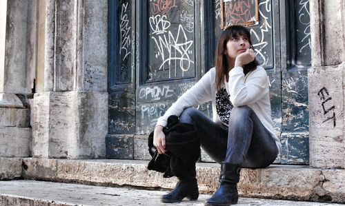 Full length of thoughtful young woman sitting on steps by graffiti door
