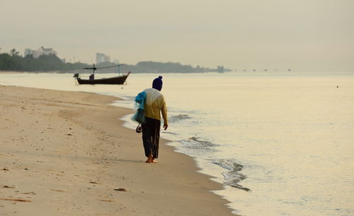 Full length rear view of fisherman holding net while walking on shore at beach