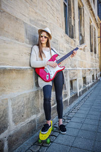 Portrait of young woman playing guitar while standing with skateboard by wall