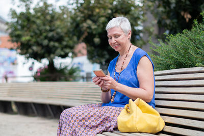 Aged woman sits on a bench in a public place in the city and uses a mobile phone