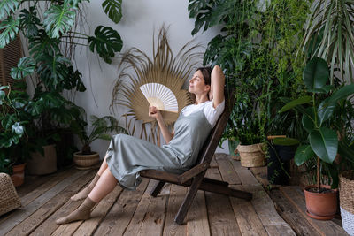 Happy woman florist refreshing in home garden breathing fresh air hold paper fan in hand at hot day