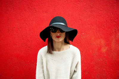 Portrait of young woman wearing sunglasses while standing against red wall