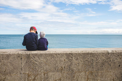 Rear view of siblings sitting on retaining wall by sea against sky