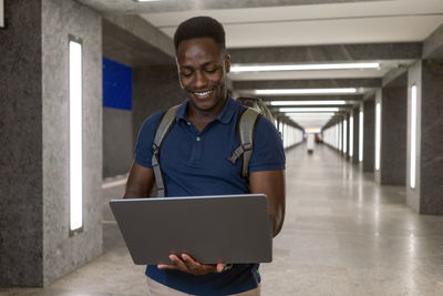 Smiling man using laptop and walking in illuminated subway tunnel