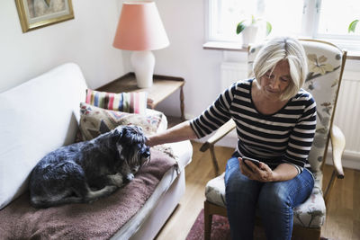 Senior woman using smart phone while stroking dog relaxing on sofa at home