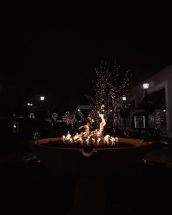 Illuminated buildings by trees against sky at night