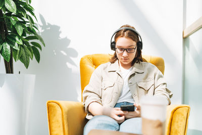 Young girl student in glasses using mobile phone, listen music in headphones in yellow chair 