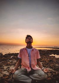 Man sitting on beach against sky during sunset