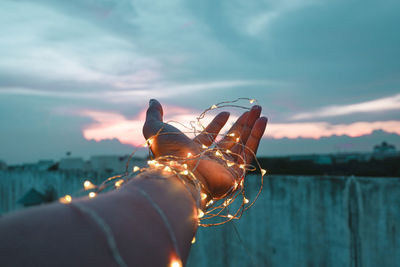 Cropped hand of person with illuminated string lights during sunset