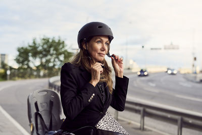 Mature businesswoman fastening helmet on street against sky