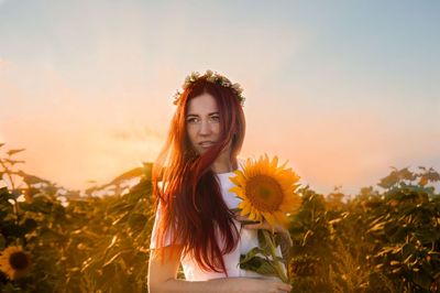 Woman holding sunflower against sky during sunset