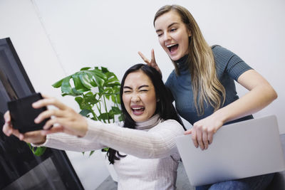 Cheerful female bloggers taking selfie through smart phone in creative office