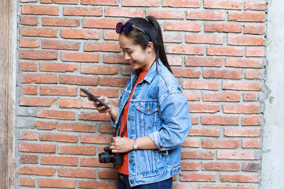Smiling woman holding phone while standing against wall 