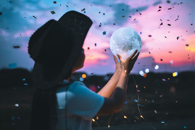 Girl holding crystal ball against sky during sunset