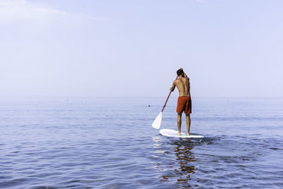 Rear view of shirtless man standing on paddleboard in sea