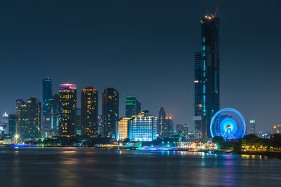 Illuminated buildings in front of chao phraya river against clear sky at night