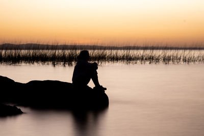 Silhouette man sitting by lake against sky during sunset