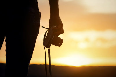 Silhouette man holding camera against sky during sunset