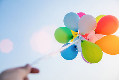 Low angle view of cropped hand holding colorful helium balloons against sky