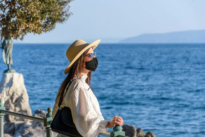 Portrait of a young woman wearing black protective mask, standing on shore of sea.