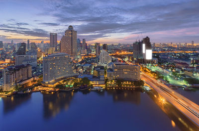 River by illuminated cityscape against sky during sunset