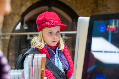 A young girl wearing a red hat sat at a computer monitor.