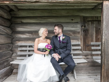 Happy bride and groom sitting on bench against wooden wall