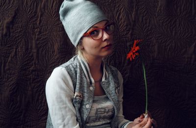 Portrait of young woman holding red gerbera daisy by brown curtain at home