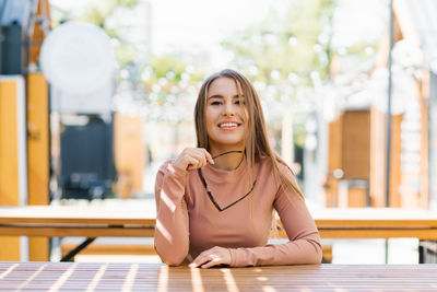 Portrait of a happy smiling woman holding sunglasses in the city in summer in a cafe and waiting 