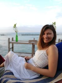 Portrait of smiling young woman sitting on chair at beach against sky