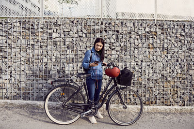 Full length of woman using smart phone while standing with bicycle against stone wall