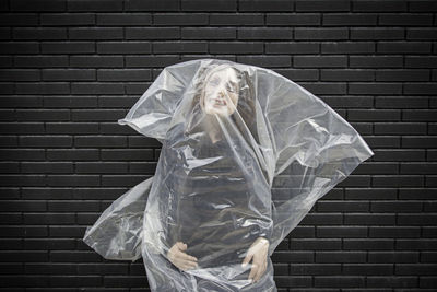 Portrait of mid adult woman in plastic bag standing against brick wall