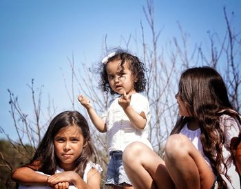 Happy friends sitting by bare tree against sky