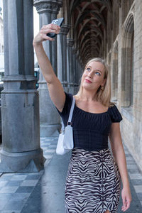 Portrait of young woman standing against historic building in a european city, taking a selfie 