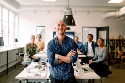 Portrait of smiling male entrepreneur standing with arms crossed and employees sitting at office desk