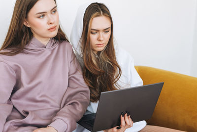 Young women sisters twins teenagers in hoodie beautiful girls using laptop sitting on yellow couch 
