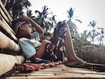 Side view of boy resting on bamboo bench against sky