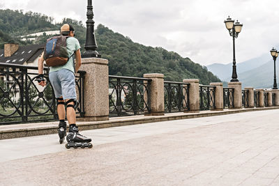 Young man in protective equipment roller skating along embankment against forest mountains in summer