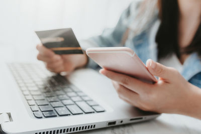 Midsection of woman holding credit card while using mobile phone and laptop at desk