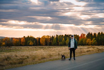 Man and french bulldog dog walking on the road against autumn forest at sunrise