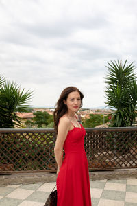 Portrait of woman wearing red dress while standing against cloudy sky