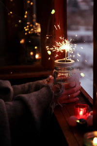Cropped hand of woman holding illuminated sparkler by window