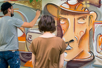 Rear view of woman looking at artist making graffiti on wall