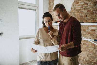 Couple discussing over blueprint in front of brick wall while renovating home